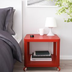 a red table with two books on it next to a white lamp and a plant