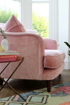 a pink velvet chair in front of a window with books on the end table next to it