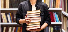 a man holding a stack of books in front of a book shelf full of books