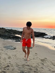 a shirtless man walking on the beach with his feet in the sand as the sun sets