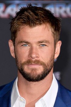 a close up of a man with a beard and blue suit jacket looking at the camera