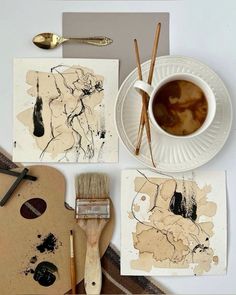 some paint and brushes are sitting on a table next to art pieces, including a cup of coffee