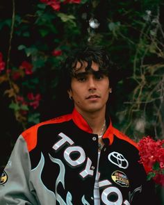 a young man in a black and grey jacket with flames on it standing next to red flowers