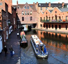 two boats traveling down a river next to tall brick buildings and people walking on the sidewalk