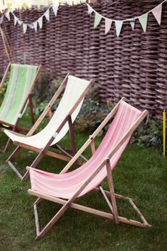 three lawn chairs sitting in the grass next to a wall with bunting flags on it