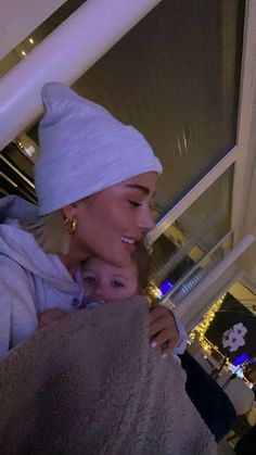 a woman holding a baby wrapped in a towel and wearing a white hat while standing next to a window