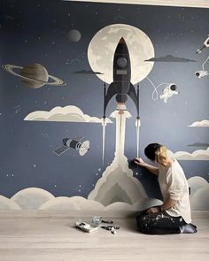 a person sitting on the floor in front of a wall painting with space and rockets