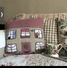 a crocheted house pillow sitting on top of a bed next to two mugs