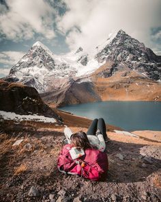 a woman laying on top of a mountain next to a body of water with snow covered mountains in the background