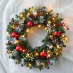 a christmas wreath with red and green ornaments on white fabric, lit up by lights