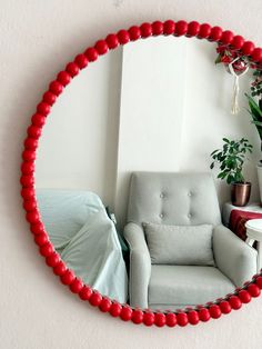 a round mirror with red beads hanging on the wall next to a chair and potted plant