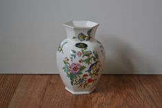 a white vase sitting on top of a wooden table next to a wall with flowers painted on it