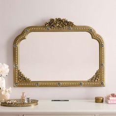 an ornate gold framed mirror above a white desk with flowers and other items on it