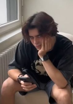 a young man sitting on a chair looking at his cell phone while holding it to his face