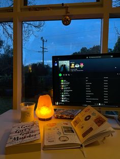 a computer monitor sitting on top of a desk next to a book and candle in front of a window