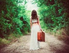 a woman walking down a dirt road with a suitcase in her hand and trees behind her