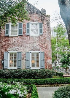 an old brick house with shutters on the windows and bushes in front of it