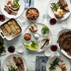 a table topped with white plates filled with meat and veggies next to glasses of wine