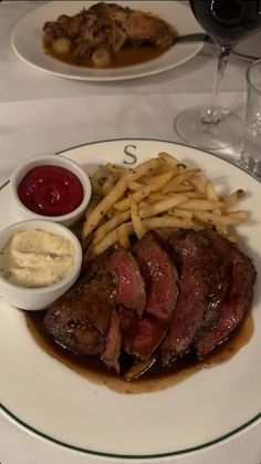 a plate with steak, fries and sauces on it next to a glass of wine