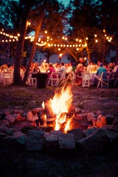 an outdoor fire pit with people sitting around it and lights strung over the trees in the background