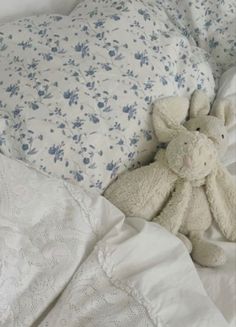 a white teddy bear laying on top of a bed next to pillows and sheets with blue flowers