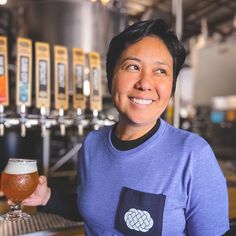 a woman holding a glass of beer in front of a row of beer kegs