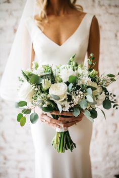 a bride holding a bouquet of white flowers and greenery in her hands with a brick wall behind her