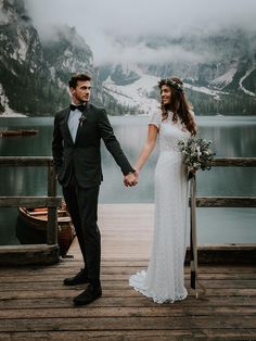 a bride and groom holding hands on a dock in front of a lake with mountains