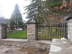 an iron fence and stone wall in front of a house with trees on the other side