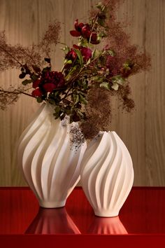 two white vases with flowers in them sitting on a red table next to a wooden wall