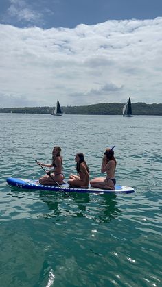 three women are sitting on a surfboard in the middle of the water with sailboats in the background