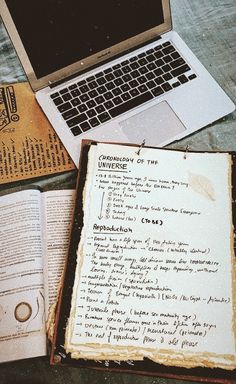 an open book sitting next to a laptop computer on top of a wooden table covered in papers