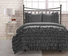 a bed with grey ruffled bedspread and pillows in front of a brick wall