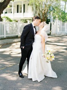 a bride and groom kissing on the street in front of a white house with trees