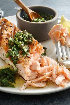 a white plate topped with salmon and greens next to a bowl of sauce on a table