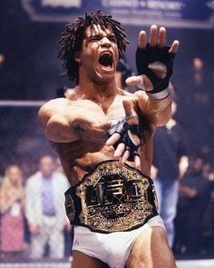 a man holding a wrestling belt in his right hand and yelling at the camera with people watching