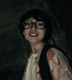 a woman with glasses and a backpack smiling at the camera while standing in front of a rock wall