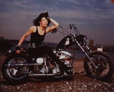 a woman sitting on top of a motorcycle next to the ocean with her hand on her head