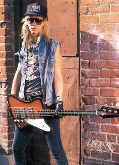 a man with long hair and sunglasses holding a guitar in front of a brick wall