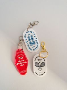 three key chains with different designs and colors on them, one has a tennis racket in it