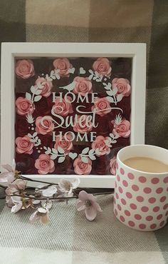 a cup of coffee sitting next to some pink flowers and a frame with the words home sweet home on it