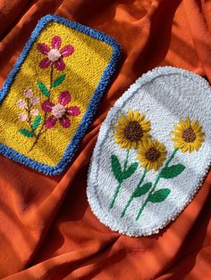 two embroidered patches with flowers on them sitting on an orange cloth covered tablecloth, next to each other