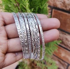 a person is holding four silver bracelets in their hand and there are plants behind them