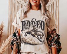 Channel the electrifying thrill of the rodeo with Lasso & Lace's "Let's Rodeo" t-shirt. This captivating design features a dynamic scene of a cowboy in full motion, expertly riding a bucking bronco, encapsulated within bold, vintage lettering that proudly declares, "Let's Rodeo". The artwork is skillfully printed on the top-tier Bella Canvas 3001 tee, renowned for its comfort and durability, making it a perfect fit for both active days and relaxed evenings. Available in three soft hues--White, N Crew Neck Letter Print Shirt For Rodeo, Western T Shirts, Bucking Bronco, Disco Cowgirl, Aesthetic Gift, Cowgirl Aesthetic, Western Graphic Tees, Rodeo Shirts, Cowgirl Shirts