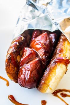 two hotdogs wrapped in tin foil with ketchup on the side and sauce drizzled around them