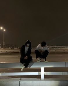 two young men sitting on the edge of an overpass at night, one wearing a mask