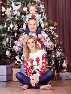 a man and woman sitting in front of a christmas tree with a baby on their shoulders