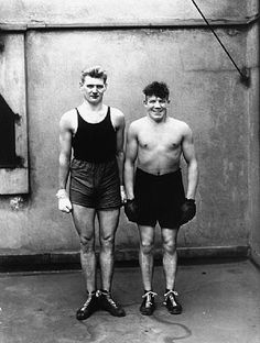 two young men standing next to each other in shorts and boxing gloves, with one holding a punching glove