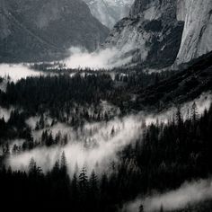 the mountains are covered in fog and clouds as they rise above them with trees on both sides