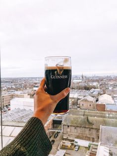 a person holding up a beer glass in front of a window overlooking a cityscape